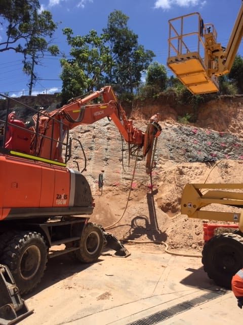 Orange Hitachi Excavator with drill mast attachment drilling horizontally into earth wall for the installation of anchors.
