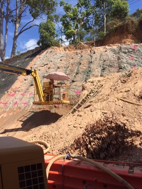 Beards team member raised up next to earth wall with the help of excavator platform, measuring and marking out position for drilling for wall anchors.