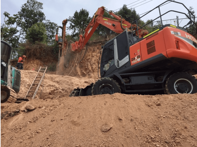 Drill mast is visible in action as orange hitachi excavator drills for holes for ground anchors for wall stabilisation.