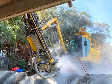 Yellow drill rig creating a dust cloud while drilling into a dam spillway for geotechnical drilling.